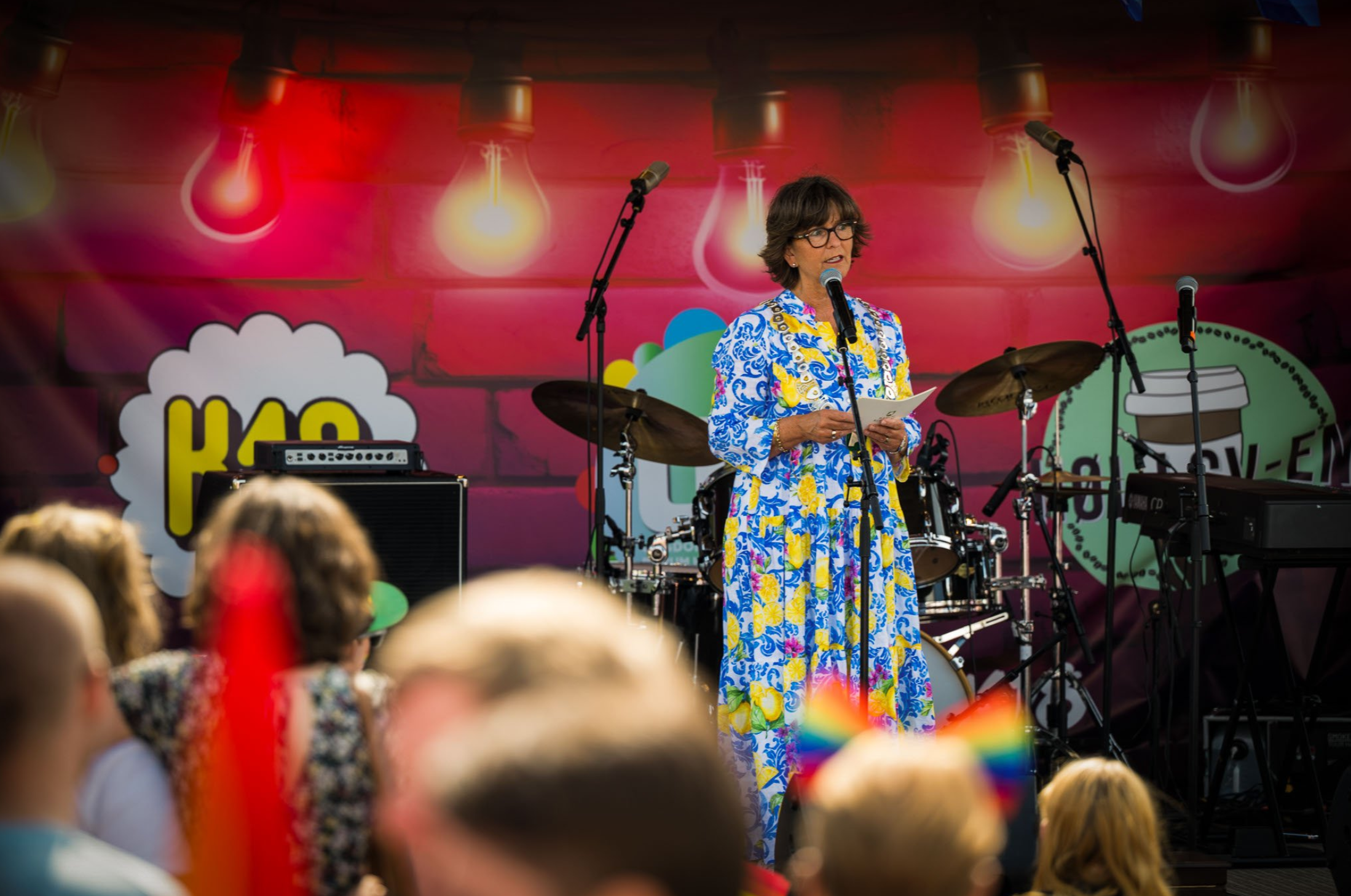 Ordfører Lisbeth Hammer Krog i Bærum står på scenen og holder tale under Sandvika pride 2025, foran et publikum med regnbueflagg og fargerik scenedekor.